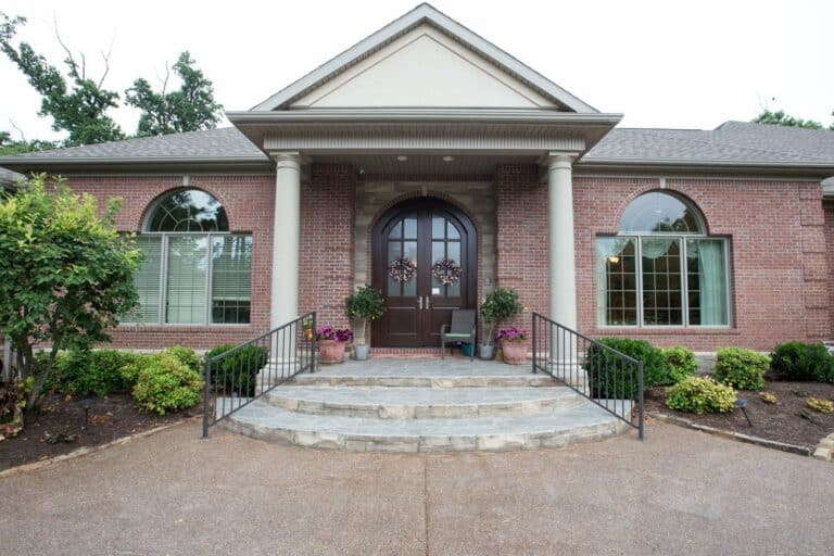 McCutchanville Custom Home Entryway Exterior - A welcoming and well-maintained front entrance of a house with excellent curb appeal. The brick facade features large arched windows on either side of the grand double front doors, which are adorned with matching decorative wreaths. Two sturdy columns support a triangular pediment above the entrance, and potted plants and flowers add a touch of greenery and color. A neatly landscaped garden frames the stone steps and iron railing, enhancing the overall attractiveness and curb appeal of the home.
