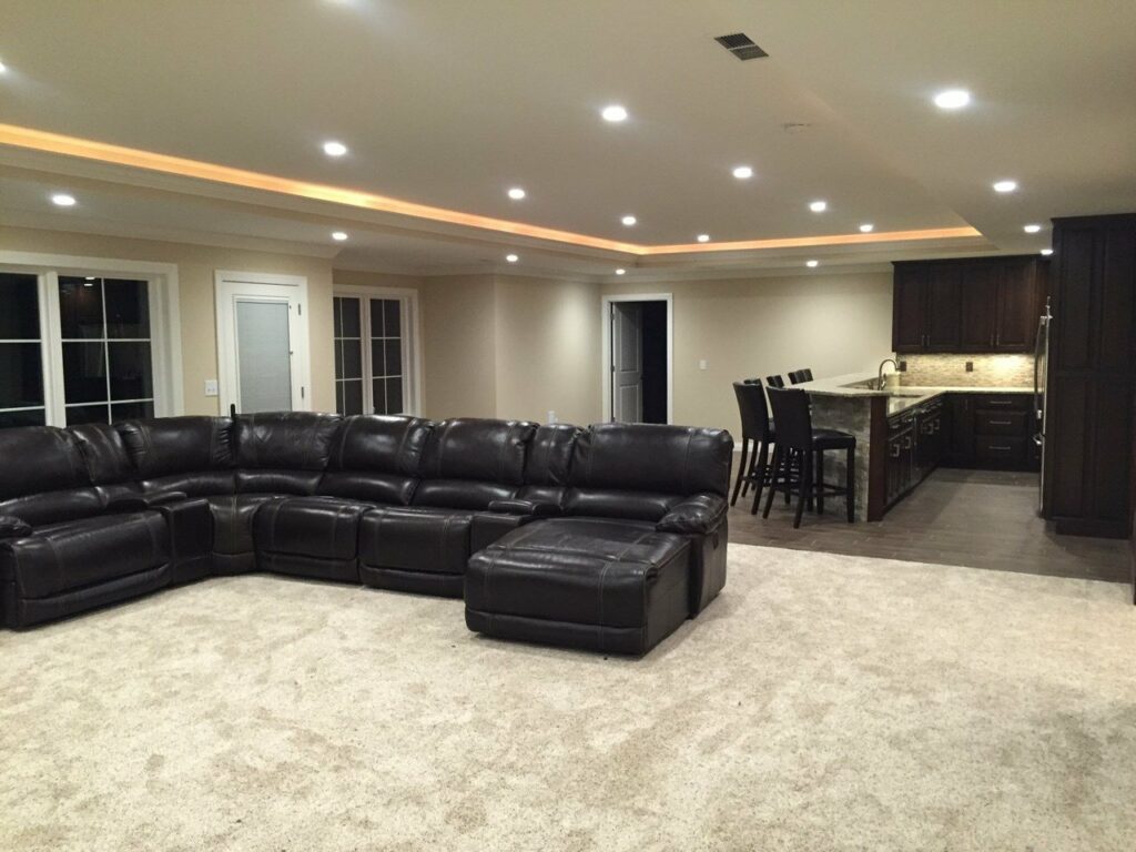 Jasper, IN Custom Home Basement - A spacious finished basement with beige walls and recessed ceiling lighting. The ceiling features an additional lighting strip around its perimeter. The room is furnished with a large, dark brown leather sectional sofa arranged in an L-shape on a light beige carpeted floor. There is an open kitchen area in the background with dark wood cabinets, a granite countertop, stainless steel appliances, and four bar stools at the counter. Multiple doors and large windows are visible, adding natural light to the room.