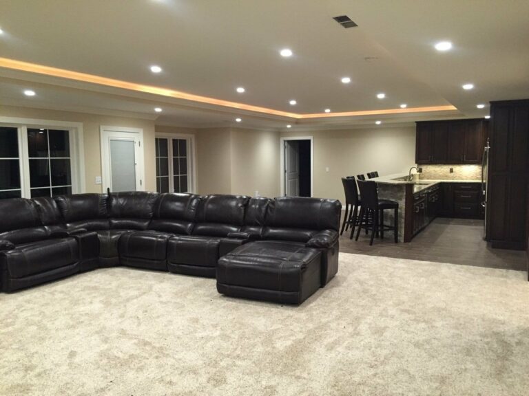 Jasper, IN Custom Home Basement - A spacious finished basement with beige walls and recessed ceiling lighting. The ceiling features an additional lighting strip around its perimeter. The room is furnished with a large, dark brown leather sectional sofa arranged in an L-shape on a light beige carpeted floor. There is an open kitchen area in the background with dark wood cabinets, a granite countertop, stainless steel appliances, and four bar stools at the counter. Multiple doors and large windows are visible, adding natural light to the room.