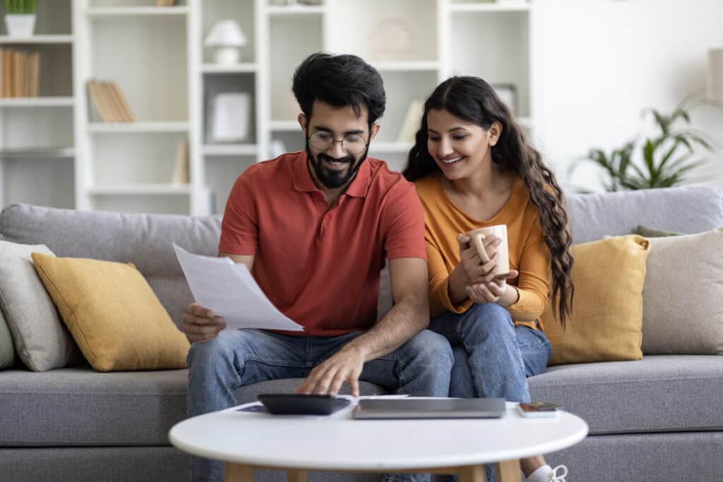 Indian Married Couple Planning Budget Together for building a home, Reading Papers And Calculating Spends While Sitting On Couch In Living Room.