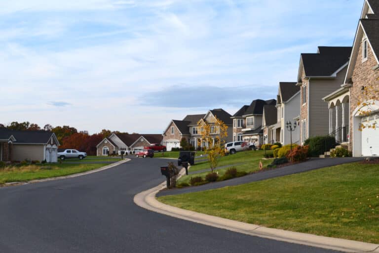 A quiet residential street featuring well-maintained houses with various architectural styles, neatly landscaped yards, and parked cars. The scene showcases a typical neighborhood under the governance of a Homeowners Association (HOA). Lush green lawns and decorative mailboxes line the curved road, reflecting a sense of community and care for property appearance.