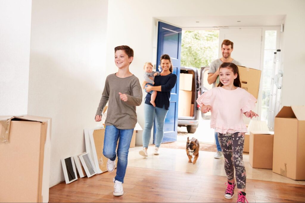 Family moving day, kids joyfully carrying boxes and playing with a dog indoors.